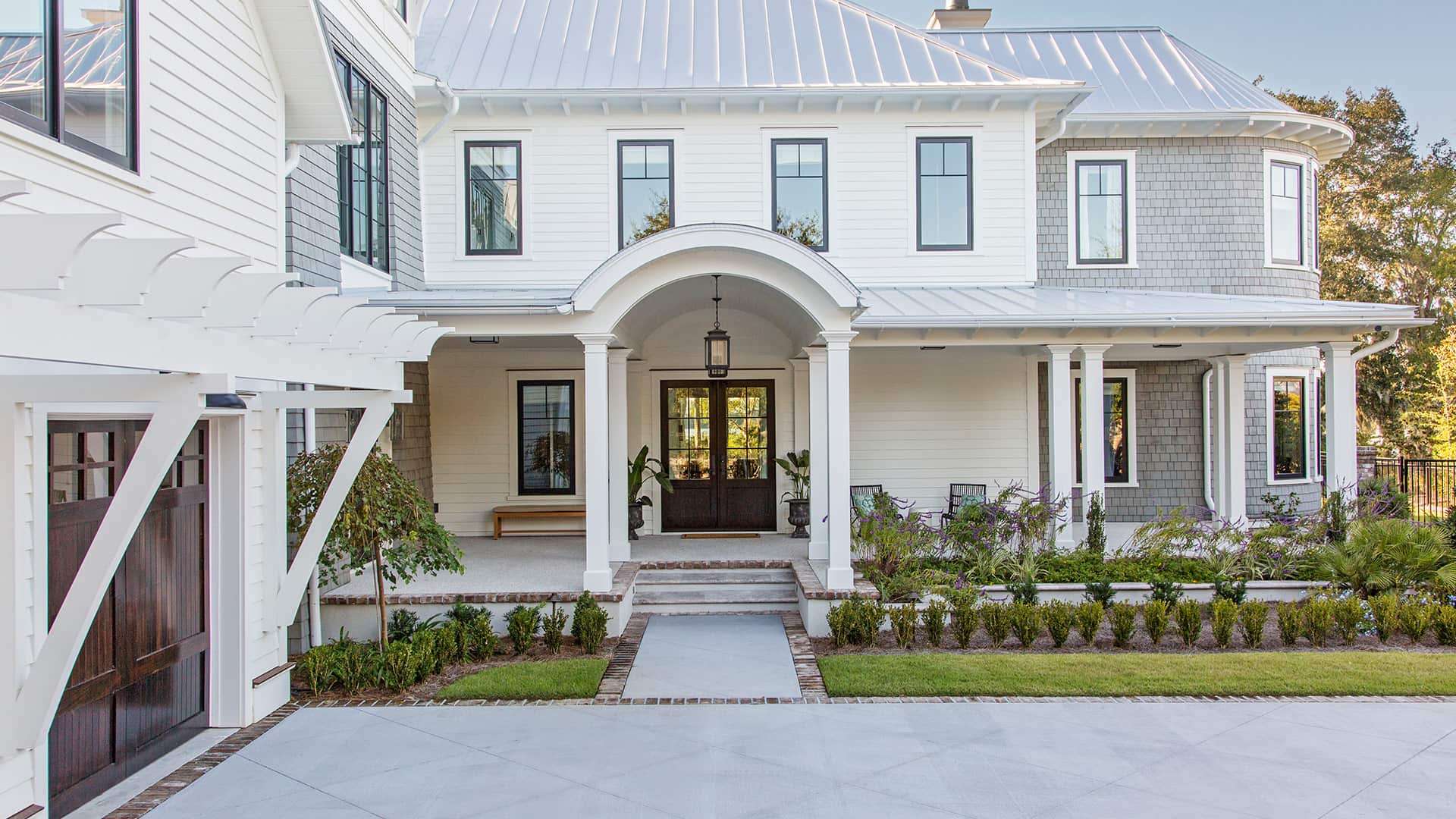 Front view of a large white coastal home with a covered porch, metal roof, and gardens.