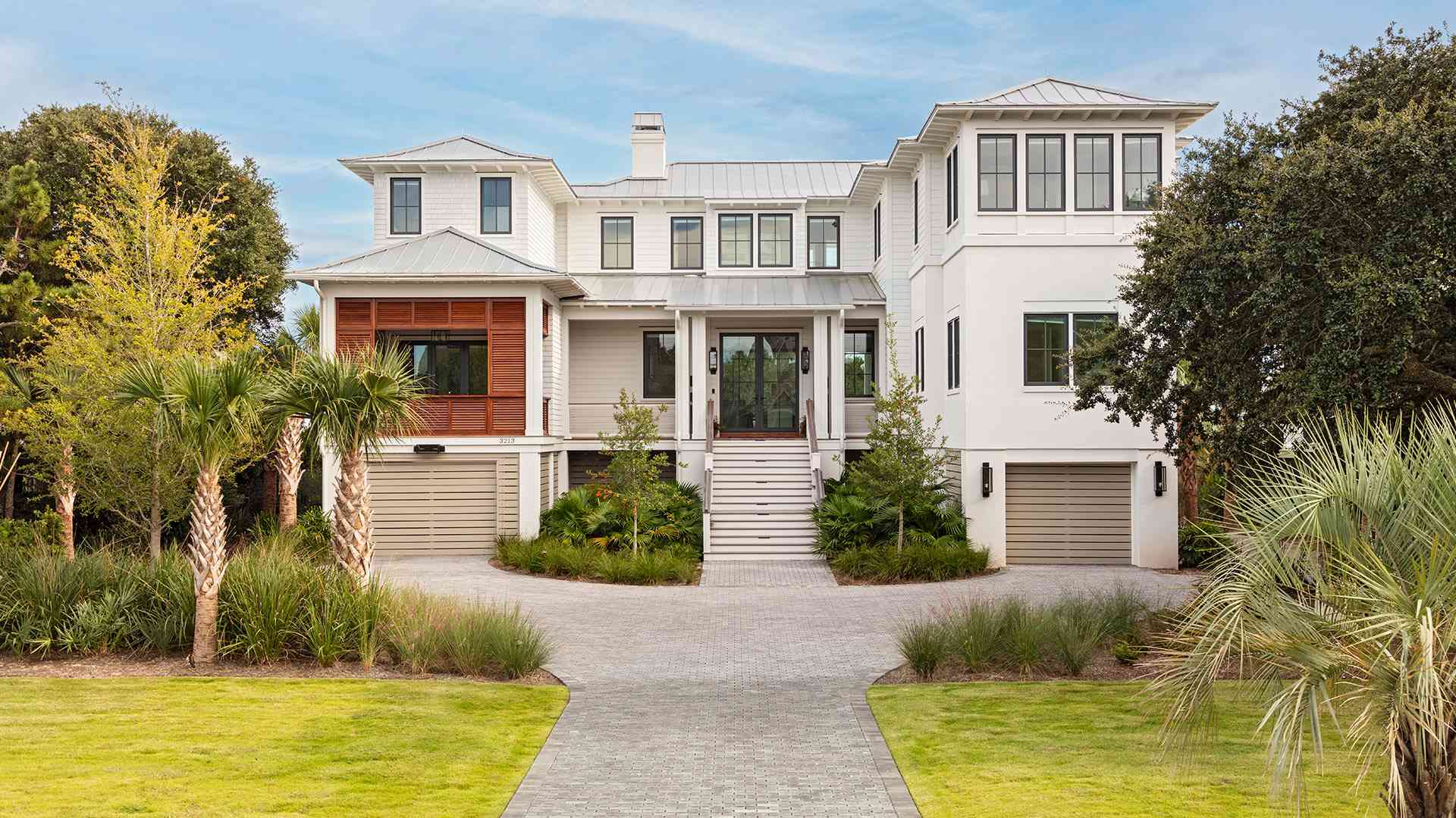 Large white coastal home with a gray metal roof, manicured lawn, palm trees, and blue sky.