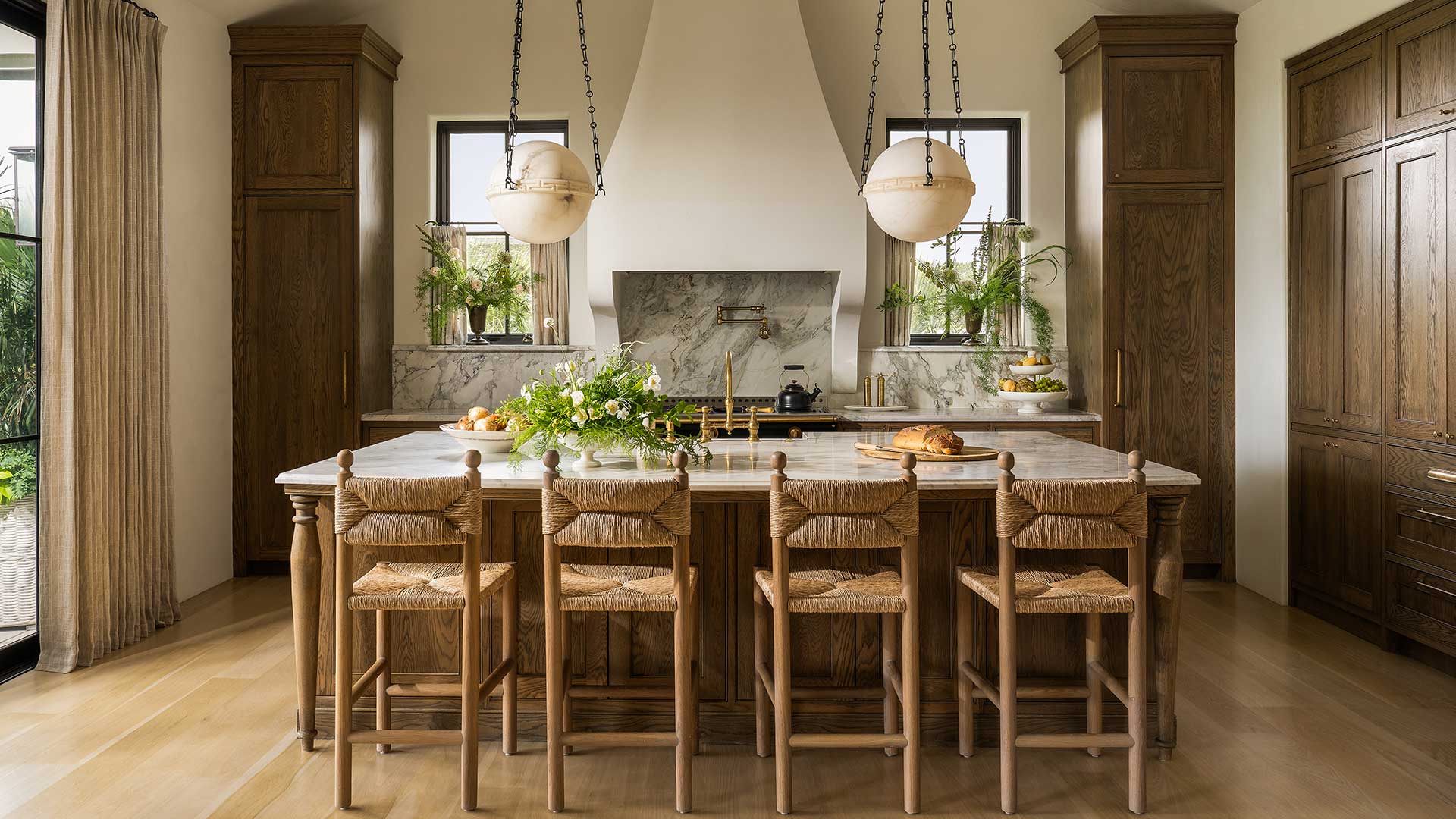 A coastal kitchen with a marble-topped island, woven rush-seat stools, alabaster globe pendants, and a plaster range hood framed by oak cabinetry.