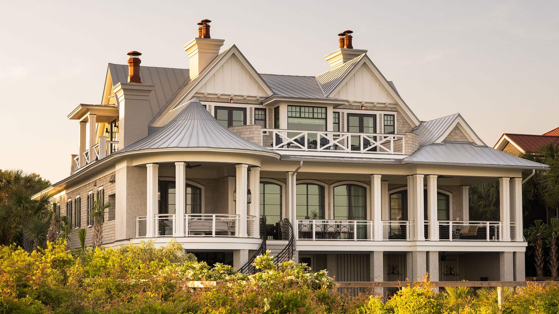 A large coastal home with a white exterior, metal roof, and multiple chimneys, surrounded by greenery.