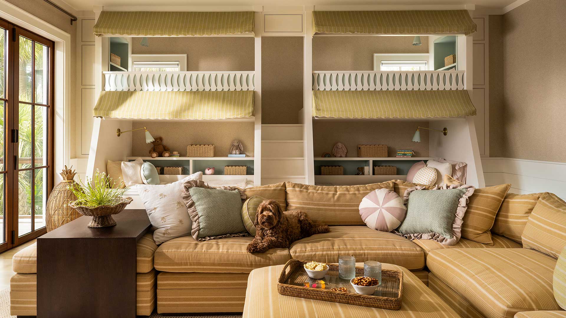 A children's bunk room with two built-in bunks under striped awnings, a wide striped sectional sofa, and a dog resting among the pillows.