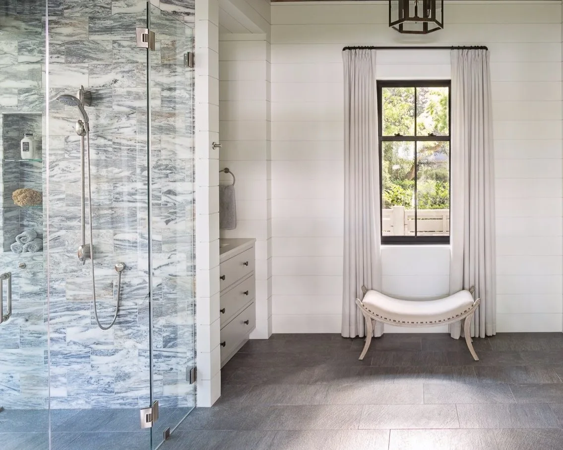 Luxury bathroom with a glass shower enclosure in gray and white marble tile and a window-side bench.