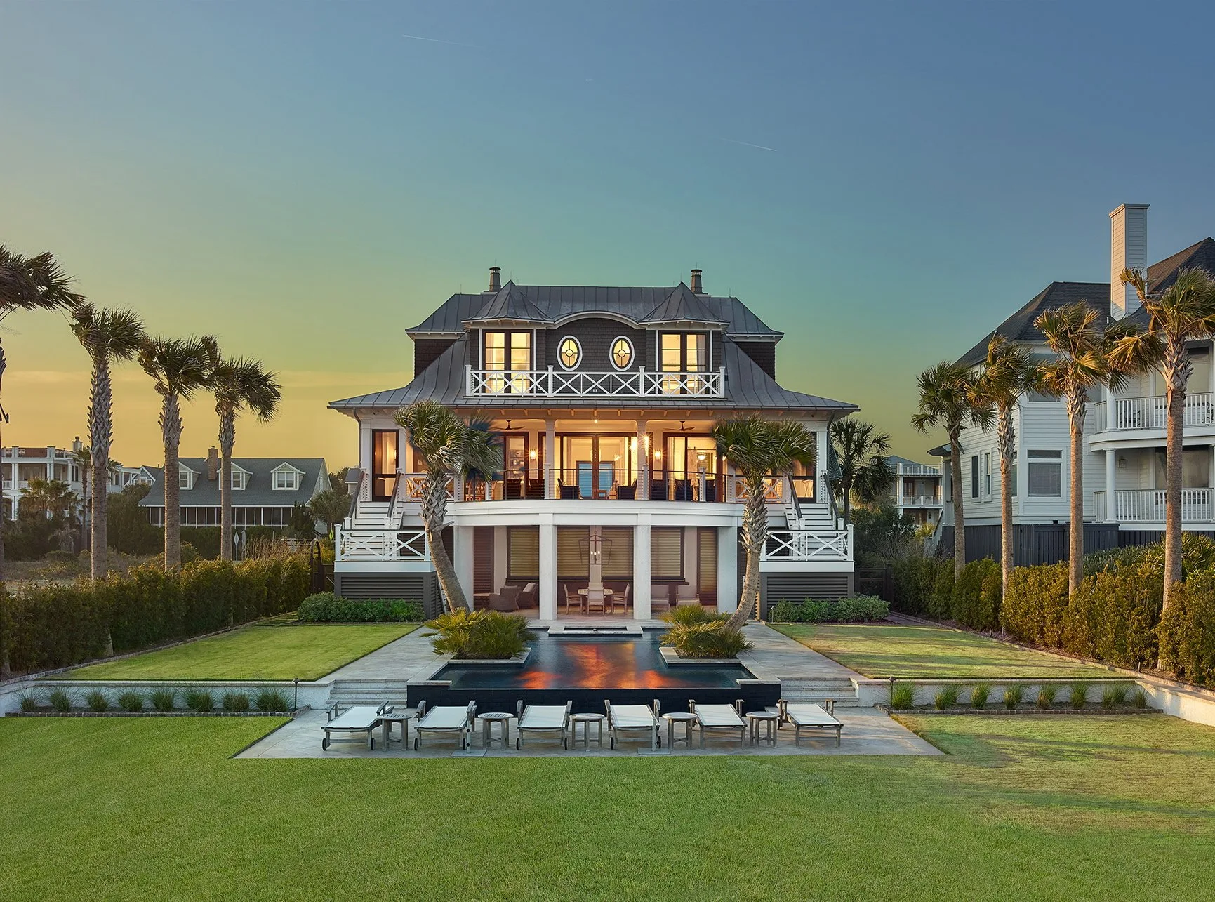 Luxury beachfront home with lawn, pool, and palm trees at sunset.