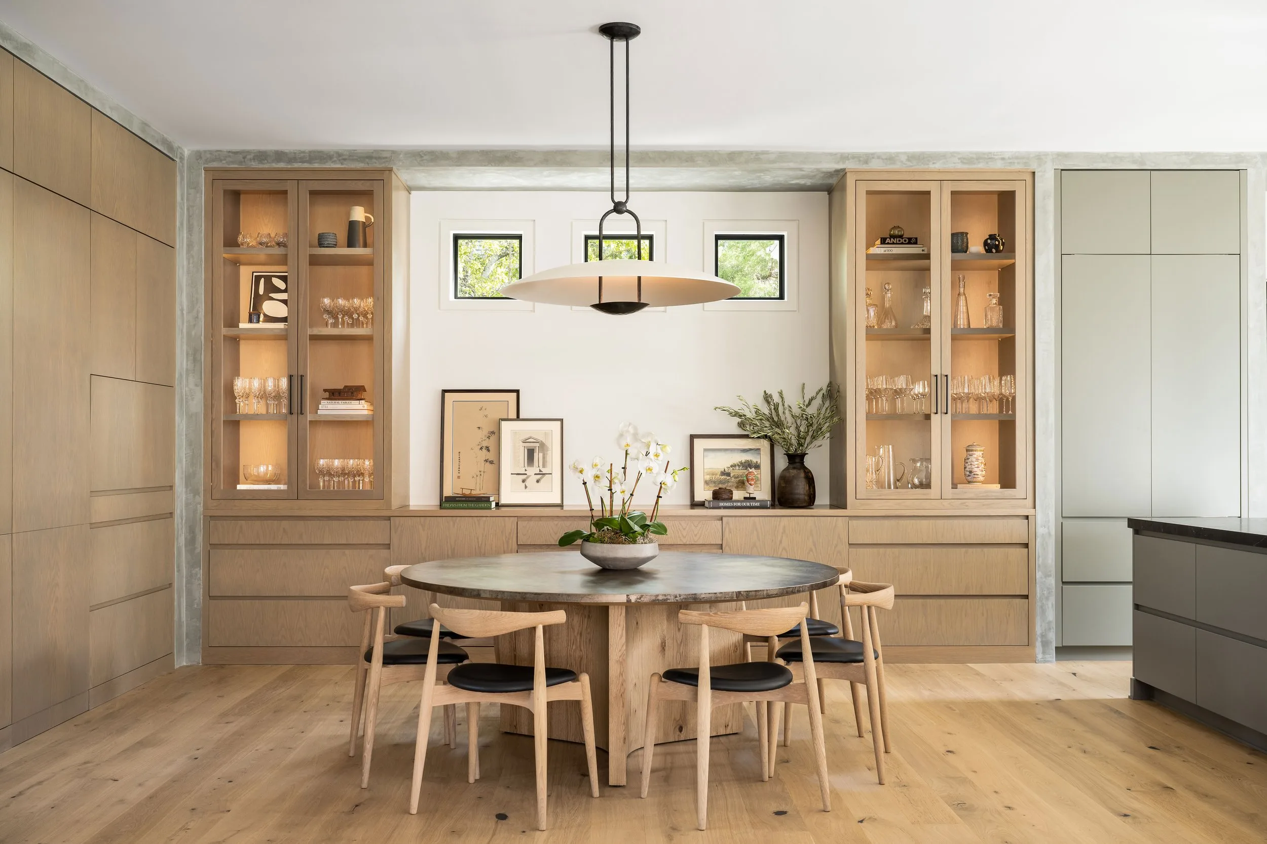 Modern dining room with a round wooden table, glass display cabinets, and a hanging light fixture.