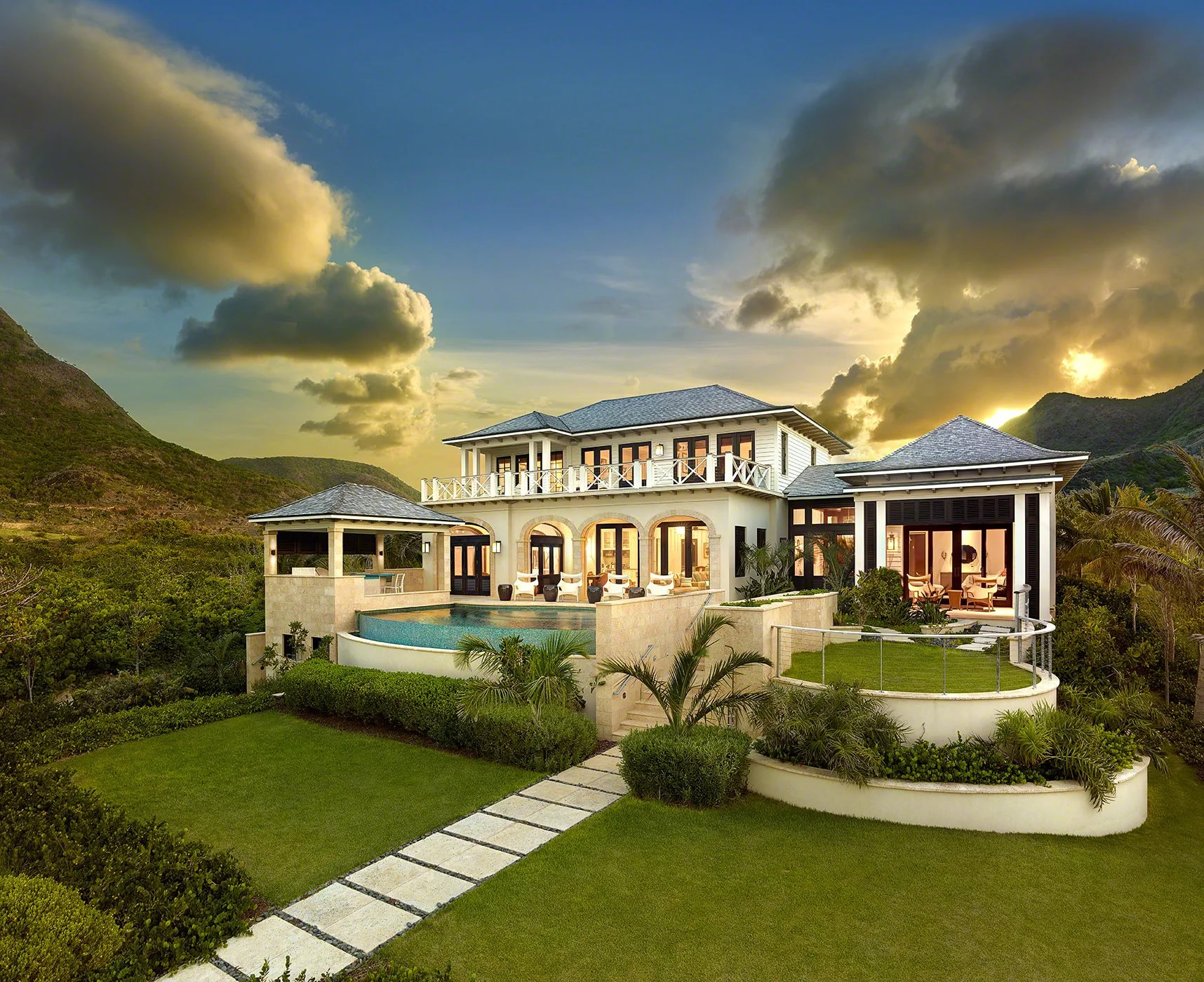 An aerial view at dusk of a Caribbean beachfront home framed by a bay and green hillsides.